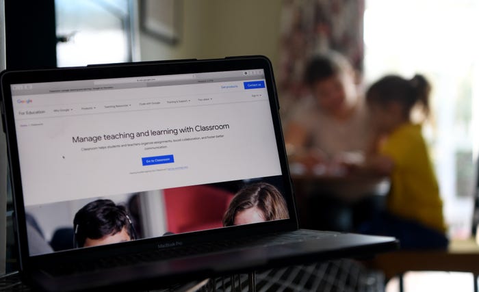 A laptop is open to Google Classroom's homepage, while a student and teacher sit in the background.