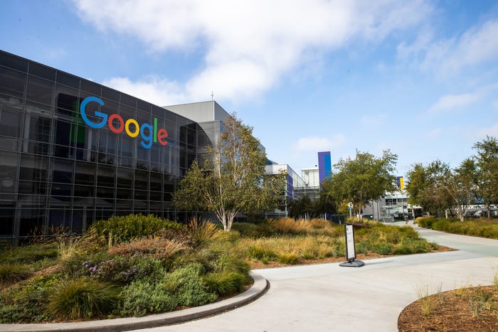 Google's sprawling headquarters in Mountain View, California, features a large Google logo on a dark glass building surrounded by trees, bushes, and pathways.