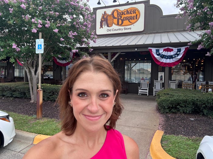 The writer taking a selfie in front of Cracker Barrel with trees with purple flowers in front of the building.