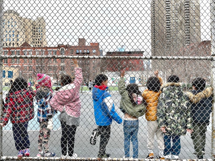 A row of students ages 5-7 raising their hands while standing along a chain-link fence on a playground during recess at P.S. 111, a public school in Hell's Kitchen, New York City.