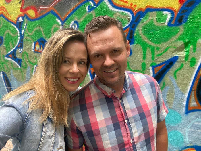 Melissa Noble and her husband sam smiling and staring at the camera while standing outside in front of a colorful mural wall.