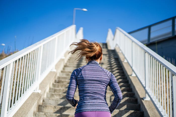 A back view of a woman with a ponytail and workout clothes prepared to go up a set of concrete stairs outside.