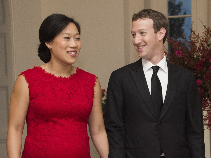 Mark Zuckerberg, wearing a black suit and tie, looking at his wife, Priscilla Chan, wearing a red lace dress, on a visit to the White House in 2015.