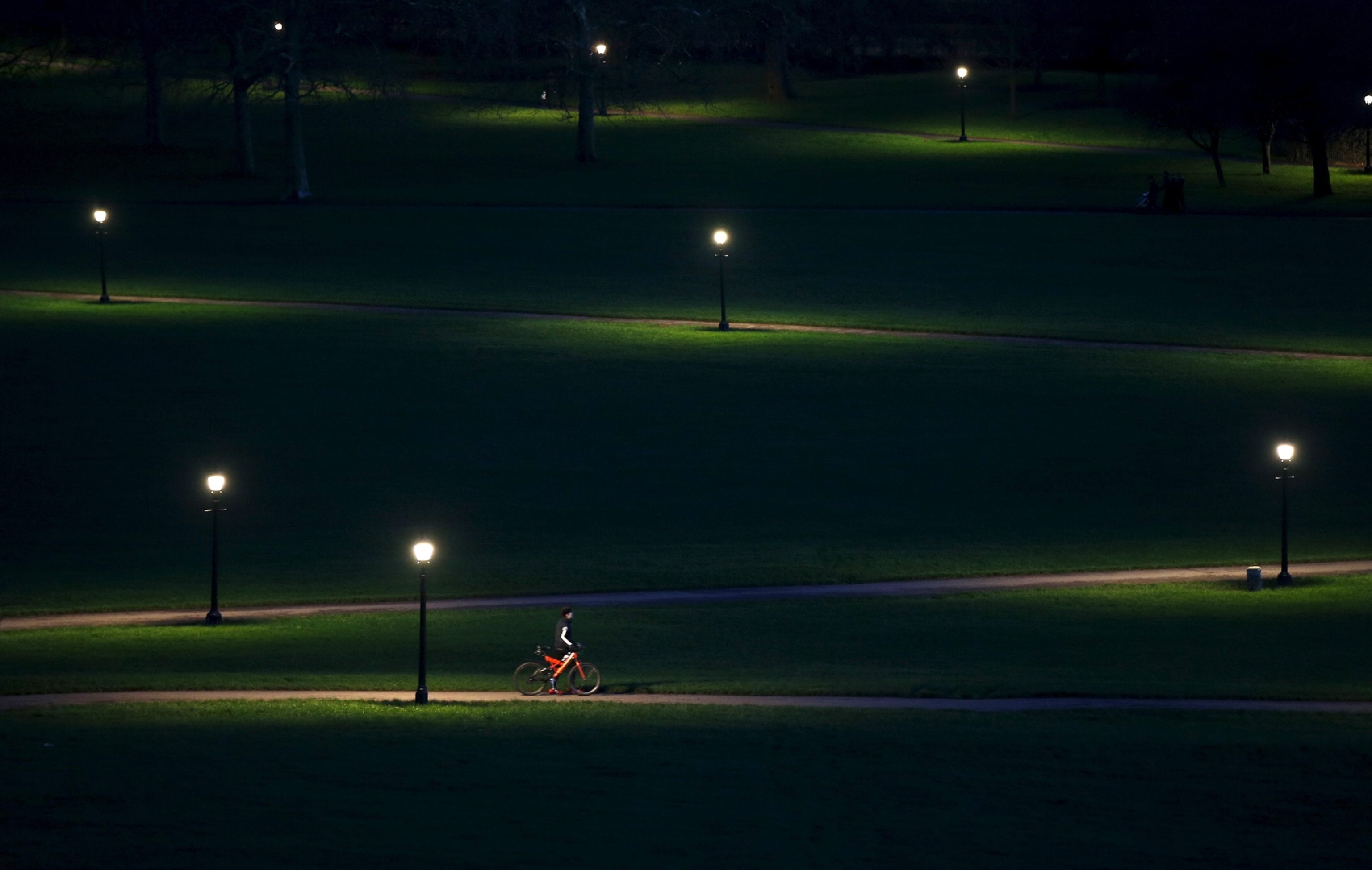 A lone cyclist riding through Primrose Hill, which is dotted with street lamps casting their light on patches of grass.