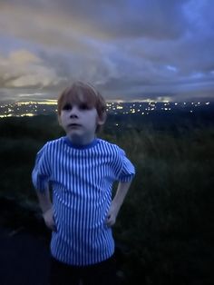 a young boy standing on top of a lush green hillside next to a city at night