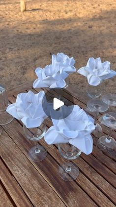 several wine glasses with white flowers in them sitting on a wooden table near the beach