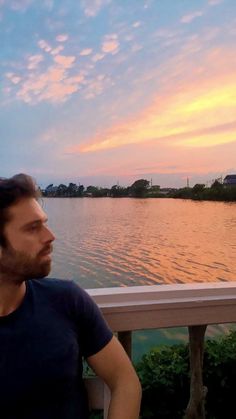 a man sitting on a bench looking out over the water at sunset with clouds in the sky