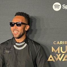 a man wearing sunglasses and a black jacket smiles at the camera while standing in front of a wall