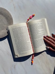 a person's hand holding an open book with beaded bracelet and hat on the table