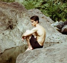 a shirtless man sitting on top of a rock next to water