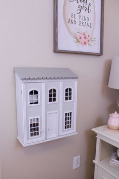 a white doll house is hanging on the wall next to a bed and nightstand with a pink lamp