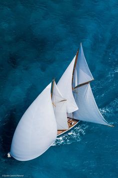 an aerial view of a sailboat in the ocean with blue water and white sails