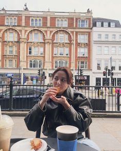 a woman sitting at an outdoor table eating pizza and drinking coffee in front of a building