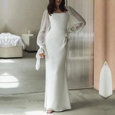 a woman standing in front of a bed wearing a white dress with sleeves on it