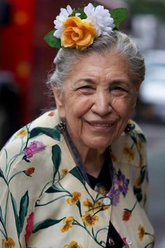 an old woman with flowers in her hair smiling at the camera while wearing a floral shirt