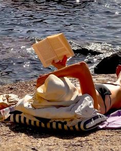 a woman laying on top of a beach next to the ocean holding an open book