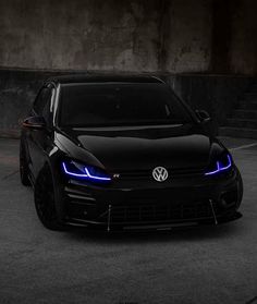 the front end of a black volkswagen car with its lights on in a parking lot