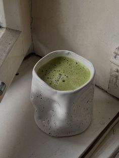 a white cup filled with green liquid sitting on top of a counter next to a window