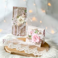 two cards with lace and flowers are on a wooden tray next to a white furnishing