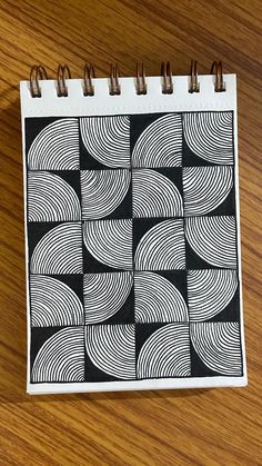 a spiral notebook with black and white designs on it sitting on a wooden table next to a cell phone