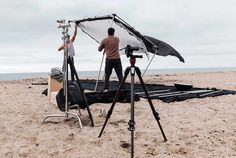 two people standing on the beach behind a camera set up to film something in the sand