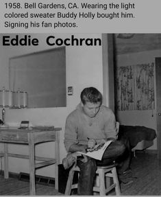 black and white photograph of man sitting on stool reading book in room with desk, chair and chalkboard