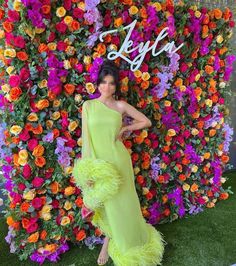 a woman standing in front of a flower wall wearing a yellow dress and ostrich feathers