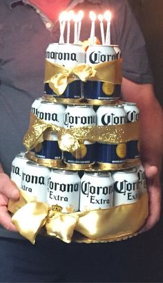 a man holding a cake made out of beer cans with candles on top and gold ribbon around it