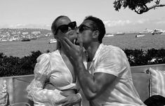 two people sitting on a bench kissing each other with the ocean and boats in the background