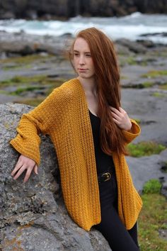 a woman sitting on top of a rock next to the ocean wearing a yellow cardigan