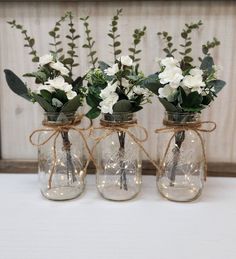 three mason jar vases with white flowers and greenery tied around the top, sitting on a table