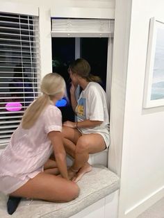 two young women sitting on a window sill talking to each other while looking out the window