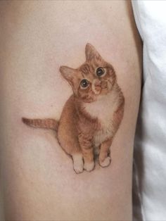 a small cat tattoo on the thigh is shown in black and grey ink, with an orange tabby kitten looking up at the viewer