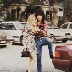 two people standing next to each other in front of some parked cars and one person is holding the woman's leg