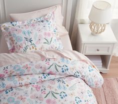 a white bed with pink and blue flowers on it, next to a night stand