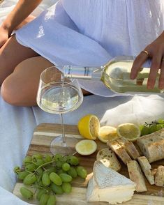 a woman pouring wine into a glass on top of a cutting board with bread and grapes