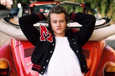 a young man is posing in front of a red car with his hands on his head