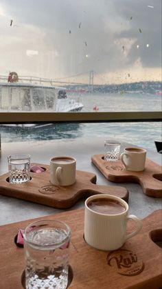 two cups of coffee sit on wooden trays near the water