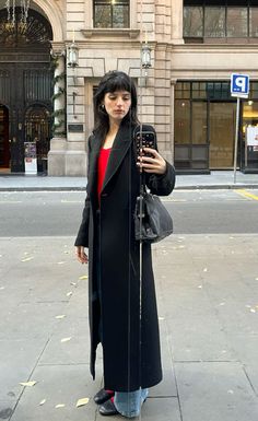 a woman is standing on the sidewalk taking a photo with her cell phone while wearing a long black coat