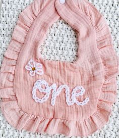 a pink bib with the word one embroidered on it, sitting on a crocheted surface