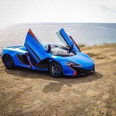 a blue sports car with its doors open sitting on the side of a road next to the ocean