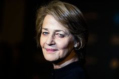 a close up of a person wearing a black suit and smiling at the camera with dark lighting in the background