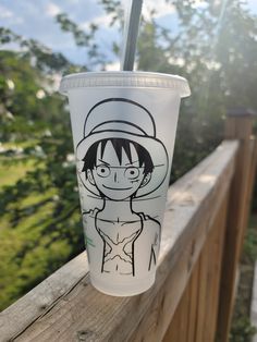 a cup with a drawing on it sitting on top of a wooden fence next to trees