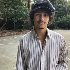 a young man wearing a hat standing in front of a parking lot with trees behind him