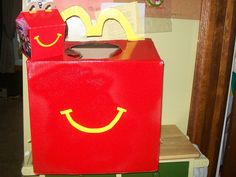 a red box with a yellow smiley face painted on the front and sides, sitting on top of a shelf