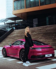 a woman standing next to a pink porsche car in front of a building with stairs