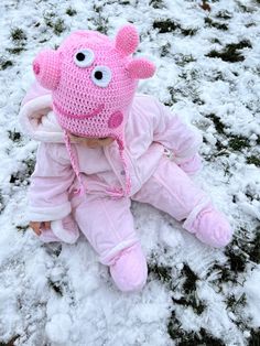 a small child wearing a pink crocheted pig hat and sitting in the snow