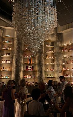 a group of people sitting at a bar with bottles on the wall behind them and a chandelier hanging from the ceiling