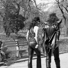 two people dressed in leathers standing next to each other near a bench and trees