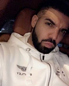 a man with a beard and white hoodie sitting in a car looking at the camera